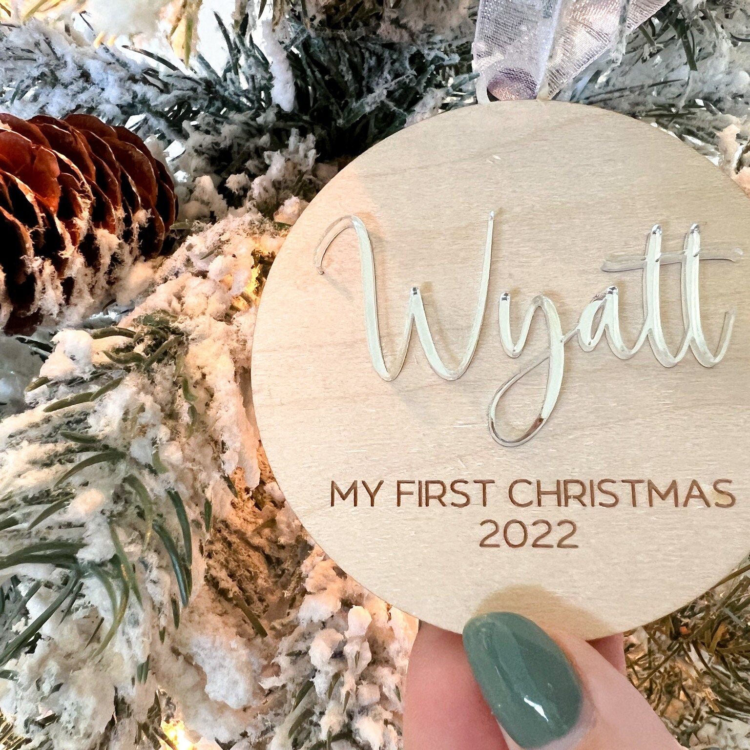 Hand holding the Baby's First Christmas Wood Ornament with a clear acrylic name against a blurred tree backdrop, ready for display.