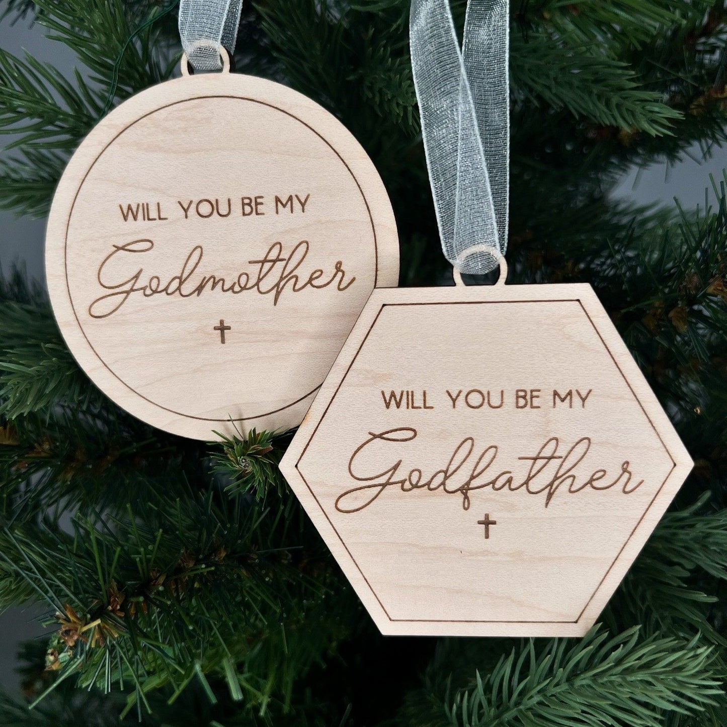 Detail shot of the script engraving on the godparent proposal ornament set, hanging on a green Christmas tree with a white sparkle ribbon.