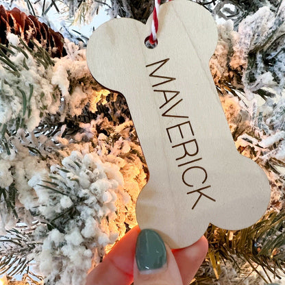 Custom Pet Name Christmas Ornament (Bone shape) being held up against a flocked Christmas tree backdrop.