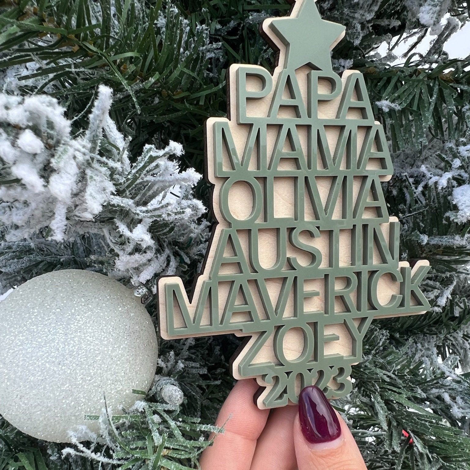 Hand holding the Family Heirloom Ornament, showcasing the thickness of the maple plywood base and the dimensional look of the 3D names.