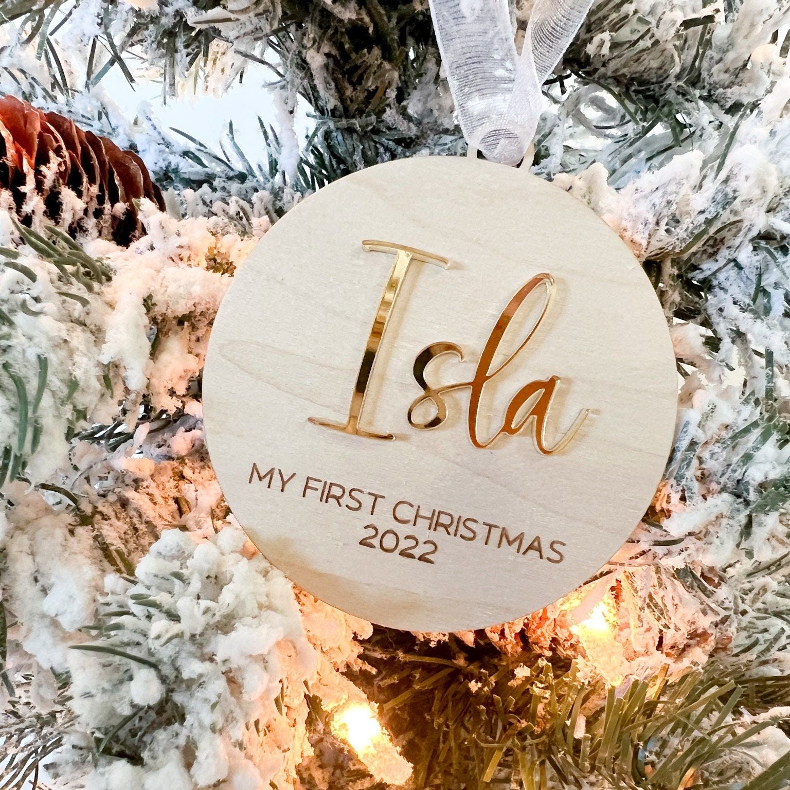 Wooden Milestone Ornament with a shiny gold mirror acrylic name ("Isla") hanging on a flocked Christmas tree.