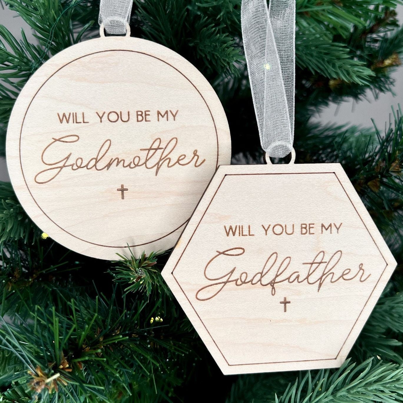 Engraved wooden Christmas ornaments with "Will You Be My Godmother" (round) and "Will You Be My Godfather" (hexagon) for a godparent proposal.