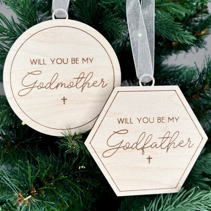 Engraved wooden Christmas ornaments with "Will You Be My Godmother" (round) and "Will You Be My Godfather" (hexagon) for a godparent proposal.