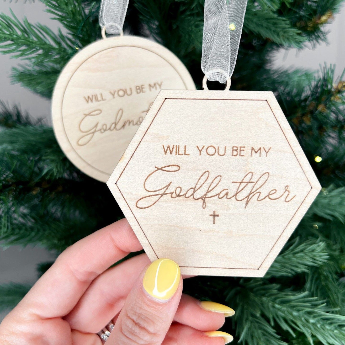 Detail shot of the script engraving on the godparent proposal ornament set, hanging on a green Christmas tree with a white sparkle ribbon.