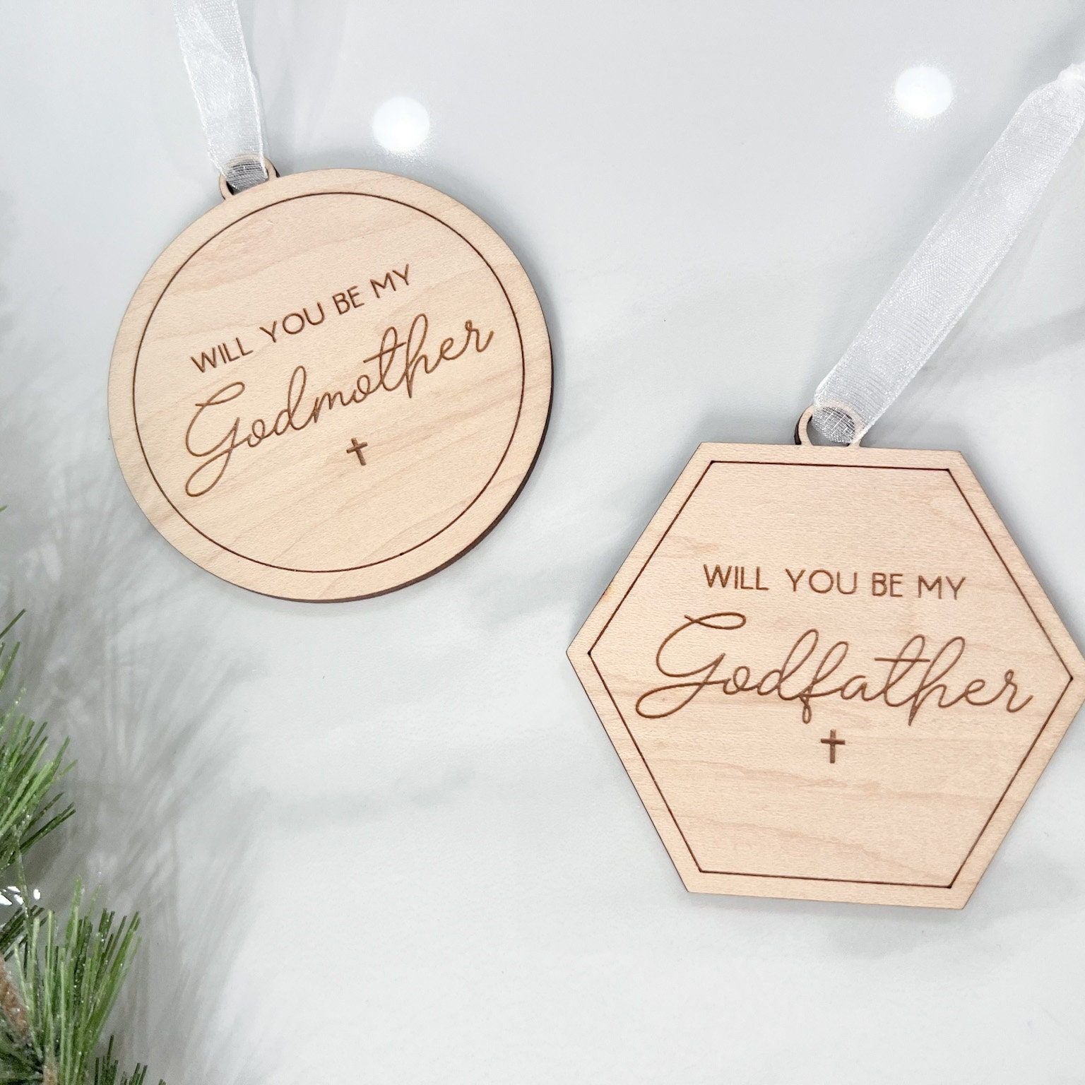 Engraved wooden Christmas ornaments with "Will You Be My Godmother" (round) and "Will You Be My Godfather" (hexagon) for a godparent proposal.