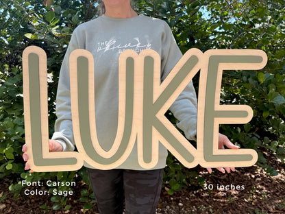 Nursery Wooden sign with baby boy name spelling "LUKE" in a Carson font with sage green and natural wood tones. The sign is 30 inches long and is held in front of a green leafy background.