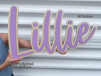 A wooden nursery name sign spelling "Lillie" in a cursive font, with a violet color and a natural wood border. The sign is 35 inches long. The font is "Delighted" and the color is violet.