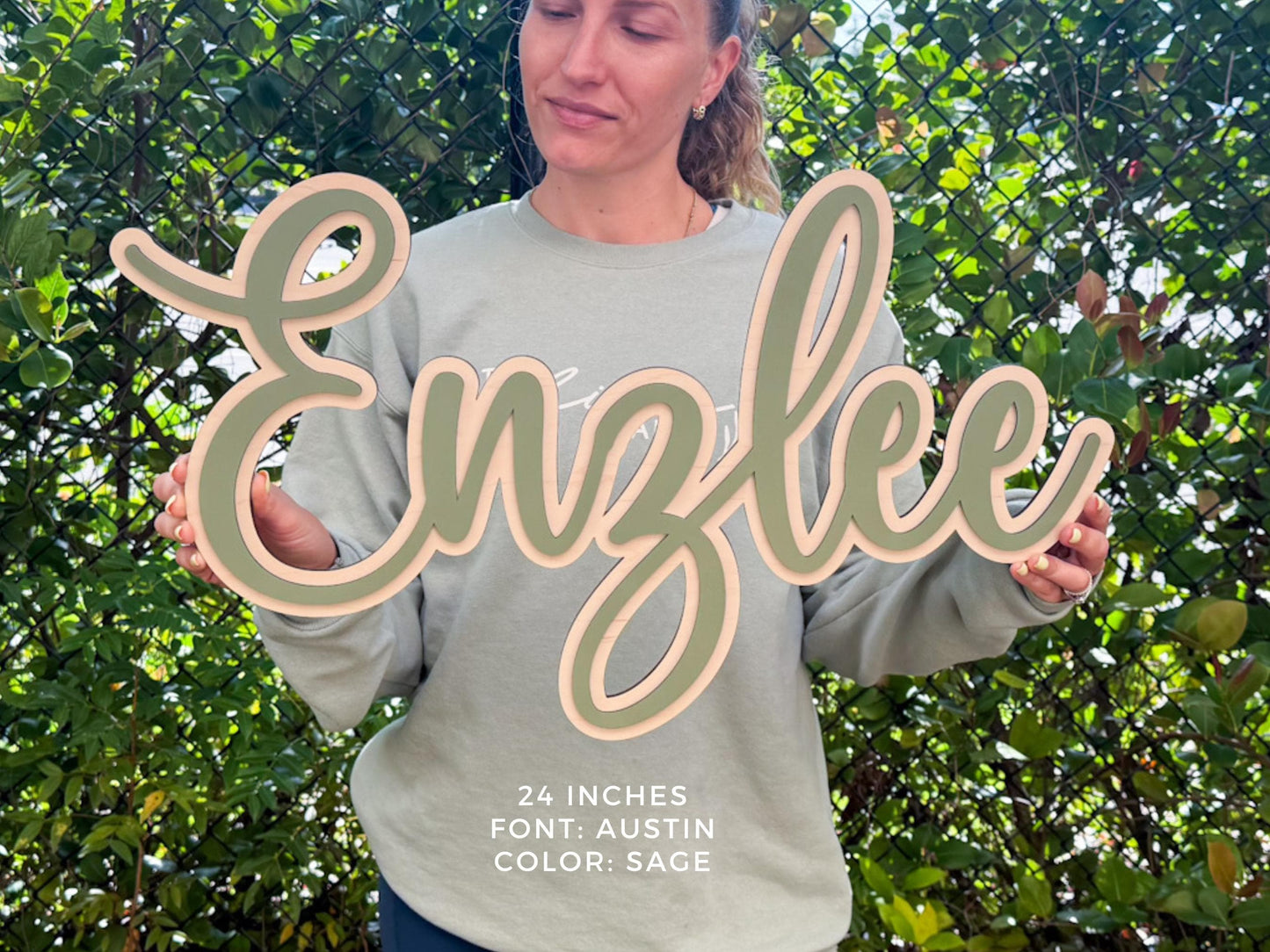 Small personalized nursery sign.  A wooden sign in the shape of the name "Enzlee" in a cursive font. The sign is 24 inches long and features a sage green color with a light pink border. The font is Austin.