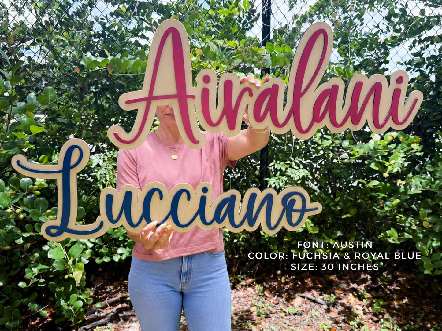 Nursery personalized baby girl wooden name signs in fuchsia and royal blue, spelling "Airvalani" and "Lucciano" in the Austin font. The signs are 30 inches in size. The names are displayed against a background of green foliage.
